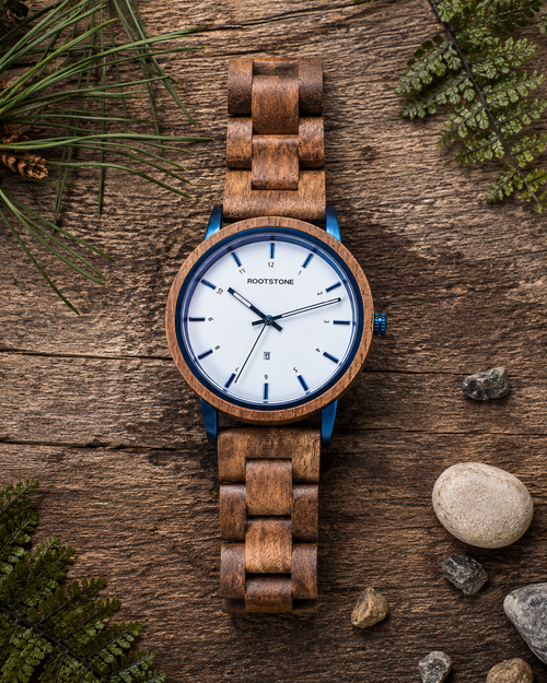 Frontalansicht der Rootstone BLÅSKOG Herrenuhr mit Holzarmband, weißem Zifferblatt und blauen Metallelementen auf rustikalem Holz